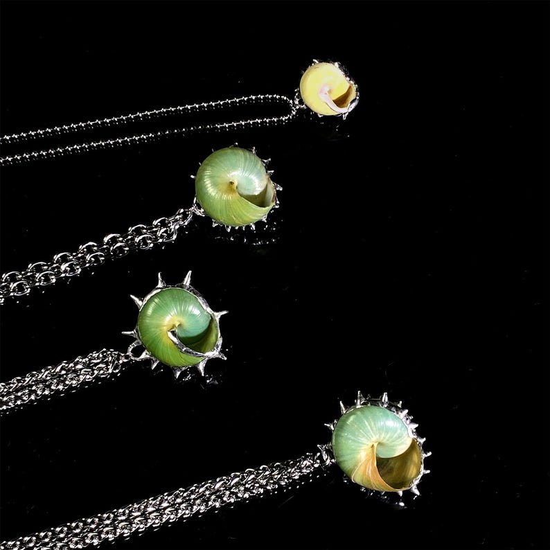 May include: Four necklaces with pendants shaped like spiral seashells. The shells are a mix of green and yellow, set in silver-colored metal. The chains are also silver-colored and have a ball chain design. The background is black.