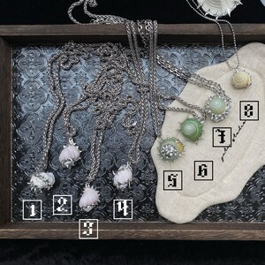 May include: A collection of silver-toned necklaces with various pendant designs. Some pendants feature light pink, green, and white gemstones, while others showcase shell-like elements. The necklaces are displayed on a dark tray with a patterned surface.