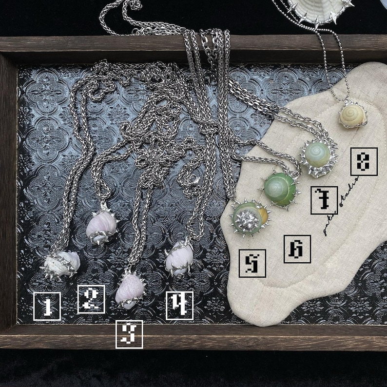 May include: A collection of silver-toned necklaces with various pendant designs. Some pendants feature light pink, green, and white gemstones, while others showcase shell-like elements. The necklaces are displayed on a dark tray with a patterned surface.