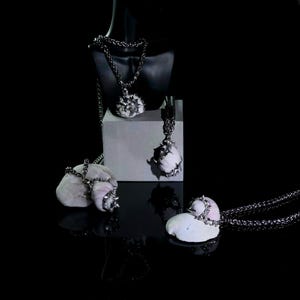 May include: A collection of unique necklaces with white, spiky pendants and silver chains. The necklaces are displayed on a black mannequin, a white block, and natural stones against a black background. The pendants have a textured appearance.