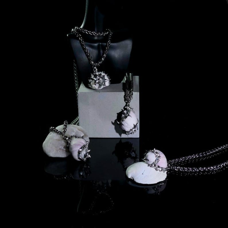 May include: A collection of unique necklaces with white, spiky pendants and silver chains. The necklaces are displayed on a black mannequin, a white block, and natural stones against a black background. The pendants have a textured appearance.