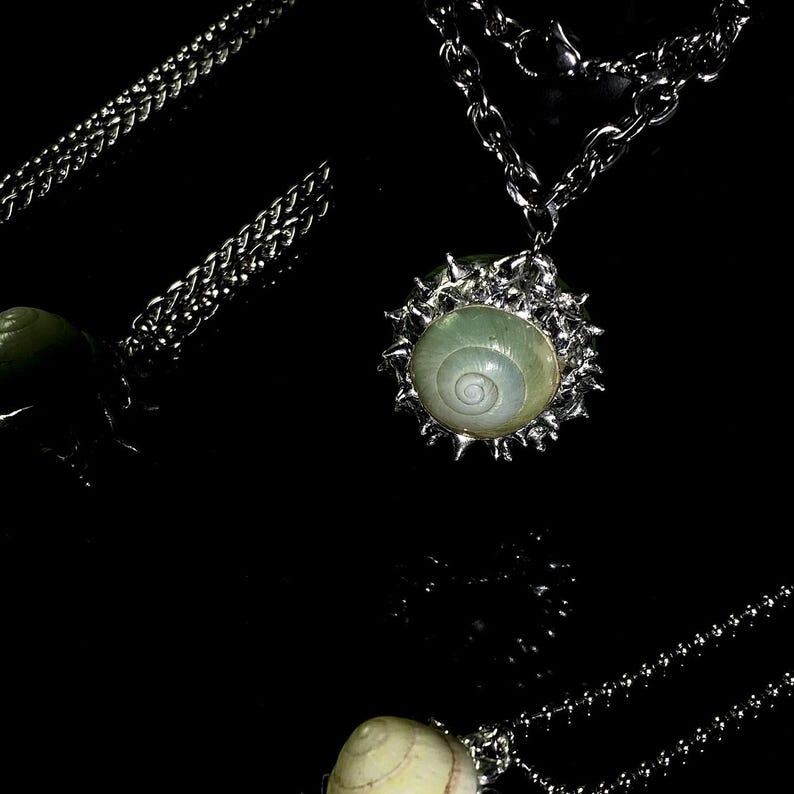 May include: A silver-toned necklace with a pendant featuring a light green, spiral-shaped shell encased in a spiky, silver-colored frame. The chain is a linked design. The background is black.