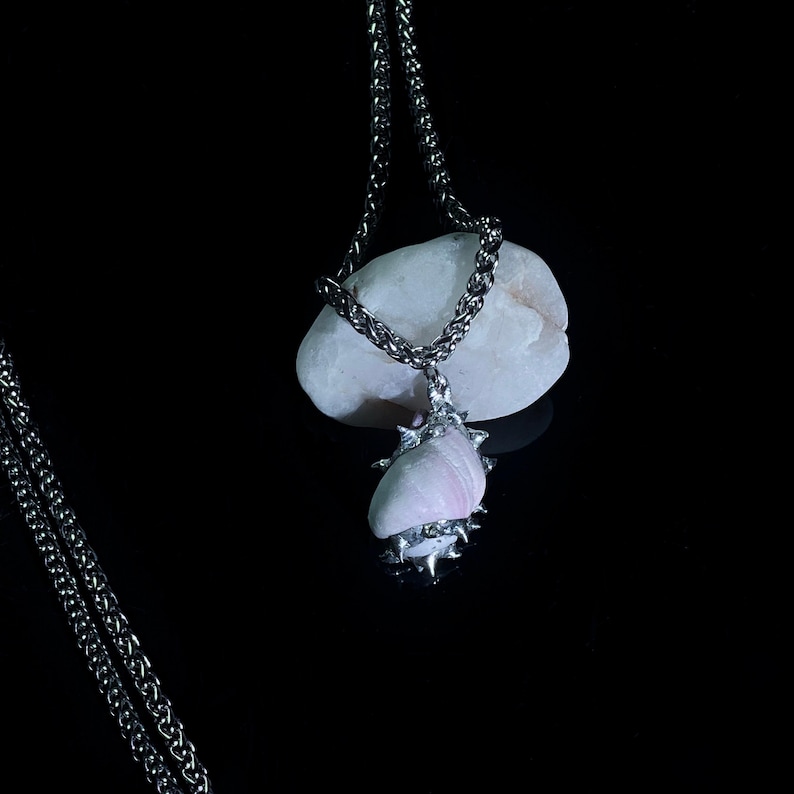 May include: A silver chain necklace with a pendant featuring a light pink and white seashell. The seashell is adorned with silver spikes and set against a black background. The necklace is displayed on a white stone.