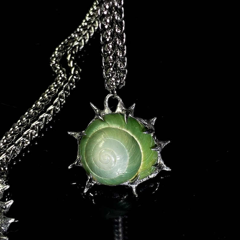 May include: A silver-toned necklace featuring a green and white spiral seashell pendant. The shell is encased in a spiky, silver-colored metal frame and hangs from a thick chain. The background is black, highlighting the jewelry.
