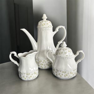 May include: A white porcelain tea set featuring a coffee pot, creamer, and sugar bowl. The set has a floral pattern in yellow and green. The coffee pot is tall with a curved spout and a lid. The creamer has a scalloped rim.
