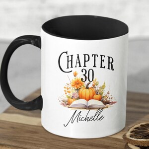 Peut inclure: Un mug en céramique blanche avec une anse et un bord intérieur noirs. L'avant présente un motif composé d'un livre ouvert, de citrouilles orange et de fleurs d'automne, avec le texte "CHAPTER 30" au-dessus et le prénom "Michelle" en dessous.
