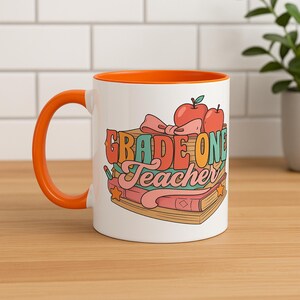 May include: White ceramic mug with an orange handle and interior. The mug features the text "GRADE ONE Teacher" in a colourful, retro design with illustrations of books, apples, and a bow. The mug is on a wooden surface.
