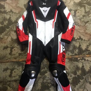 Peut inclure: Combinaison de course de moto noire, blanche et rouge. Le nom de la marque "DAINESE" est visible sur les manches. La combinaison a un panneau avant blanc, des accents rouges et des protections aux genoux. Le nom "Dair" est imprim&eacute; sur la poitrine.