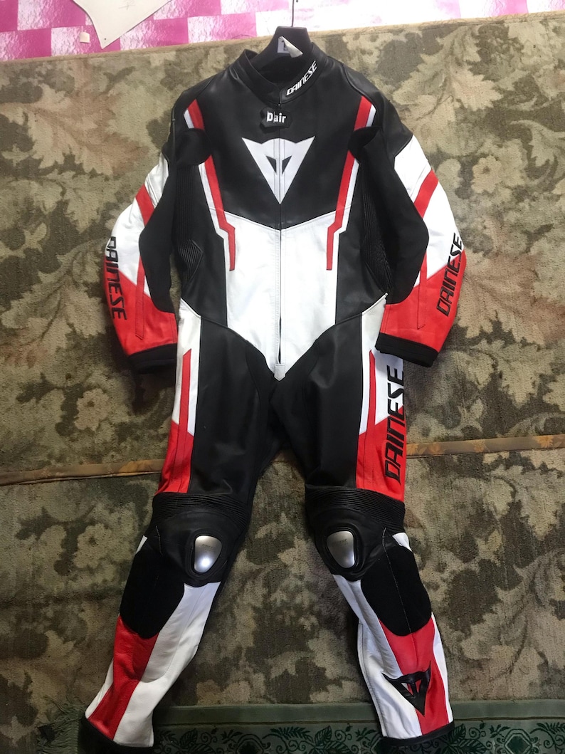 Peut inclure: Combinaison de course de moto noire, blanche et rouge. Le nom de la marque "DAINESE" est visible sur les manches. La combinaison a un panneau avant blanc, des accents rouges et des protections aux genoux. Le nom "Dair" est imprim&eacute; sur la poitrine.