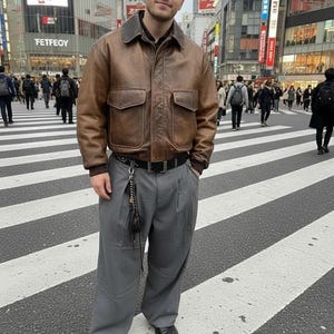 Puede incluir: Una persona con una chaqueta de cuero marrón con dos bolsillos delanteros, un cinturón negro y pantalones grises con pliegues. La persona está de pie en un paso de cebra en una ciudad con edificios y letreros al fondo.