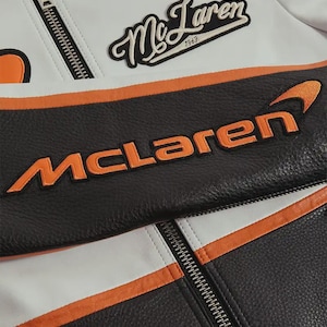 May include: Close-up of a black, white, and orange McLaren racing jacket. The jacket features the McLaren logo in orange and the McLaren name embroidered in orange. The jacket has a zipper and a patch that says "McLaren 1963".
