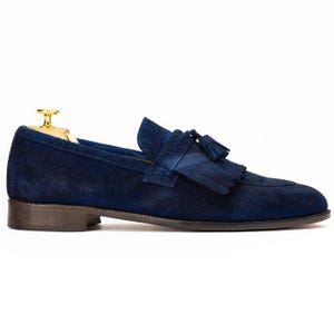 Puede incluir: Mocas&iacute;n de ante azul marino con detalle de borlas. El zapato tiene una puntera redondeada y una suela marr&oacute;n oscuro. Se inserta una horma dorada dentro del zapato.