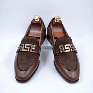 K&ouml;nnte beinhalten: Ein Paar braune Leder-Loafer mit goldfarbener Schnalle. Die Schuhe haben einen Wildleder-Akzent am Blatt und werden mit h&ouml;lzernen Schuhspannern pr&auml;sentiert. Das Leder hat eine strukturierte Optik.