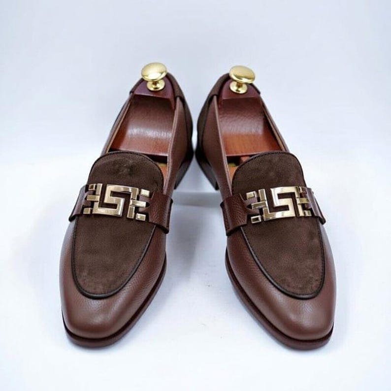 K&ouml;nnte beinhalten: Ein Paar braune Leder-Loafer mit goldfarbener Schnalle. Die Schuhe haben einen Wildleder-Akzent am Blatt und werden mit h&ouml;lzernen Schuhspannern pr&auml;sentiert. Das Leder hat eine strukturierte Optik.