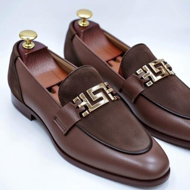 K&ouml;nnte beinhalten: Ein Paar braune Leder-Loafer mit einer goldfarbenen Zierschnalle. Die Schuhe haben einen Wildlederakzent auf dem Fu&szlig;r&uuml;cken und werden auf einer wei&szlig;en Oberfl&auml;che pr&auml;sentiert. Die Schuhe sind neu und ungetragen.