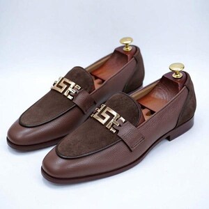 K&ouml;nnte beinhalten: Ein Paar braune Leder-Loafer mit Wildleder-Akzenten und goldfarbenen Metallschnallen. Die Schuhe werden auf Schuhspannern aus Holz pr&auml;sentiert. Die Loafer haben ein klassisches Design f&uuml;r formelle oder halbformelle Anl&auml;sse.