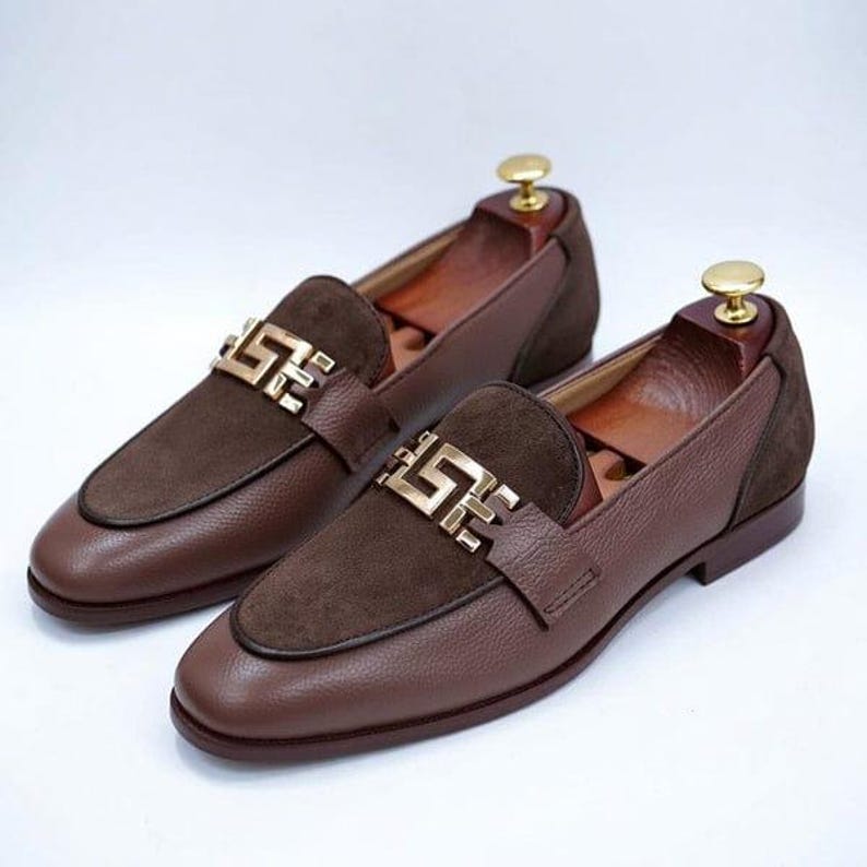 K&ouml;nnte beinhalten: Ein Paar braune Leder-Loafer mit Wildleder-Akzenten und goldfarbenen Metallschnallen. Die Schuhe werden auf Schuhspannern aus Holz pr&auml;sentiert. Die Loafer haben ein klassisches Design f&uuml;r formelle oder halbformelle Anl&auml;sse.
