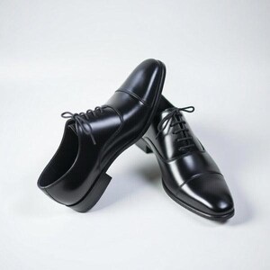 May include: A pair of black leather oxford shoes featuring a cap toe design, a low block heel, and thin black laces. The shoes have a smooth, polished finish and a structured silhouette with a rounded toe box, presented against a plain background.