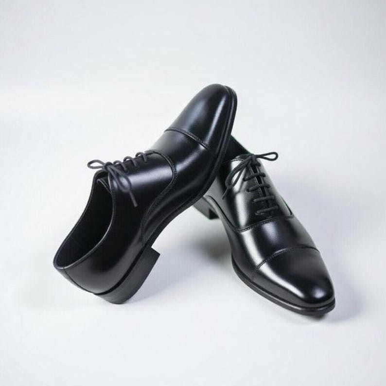 May include: A pair of black leather oxford shoes featuring a cap toe design, a low block heel, and thin black laces. The shoes have a smooth, polished finish and a structured silhouette with a rounded toe box, presented against a plain background.