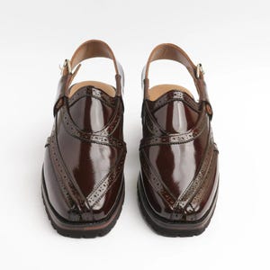 May include: A pair of brown leather shoes with a glossy finish. The shoes have a strap around the ankle and decorative perforations along the edges. The soles are dark brown, and the shoes appear to be new.