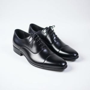 May include: A pair of black leather oxford shoes featuring a cap toe design, a closed lacing system with thin black laces, and a low heel. The shoes have a smooth, polished finish and a rounded toe shape, presenting a clean and structured appearance.