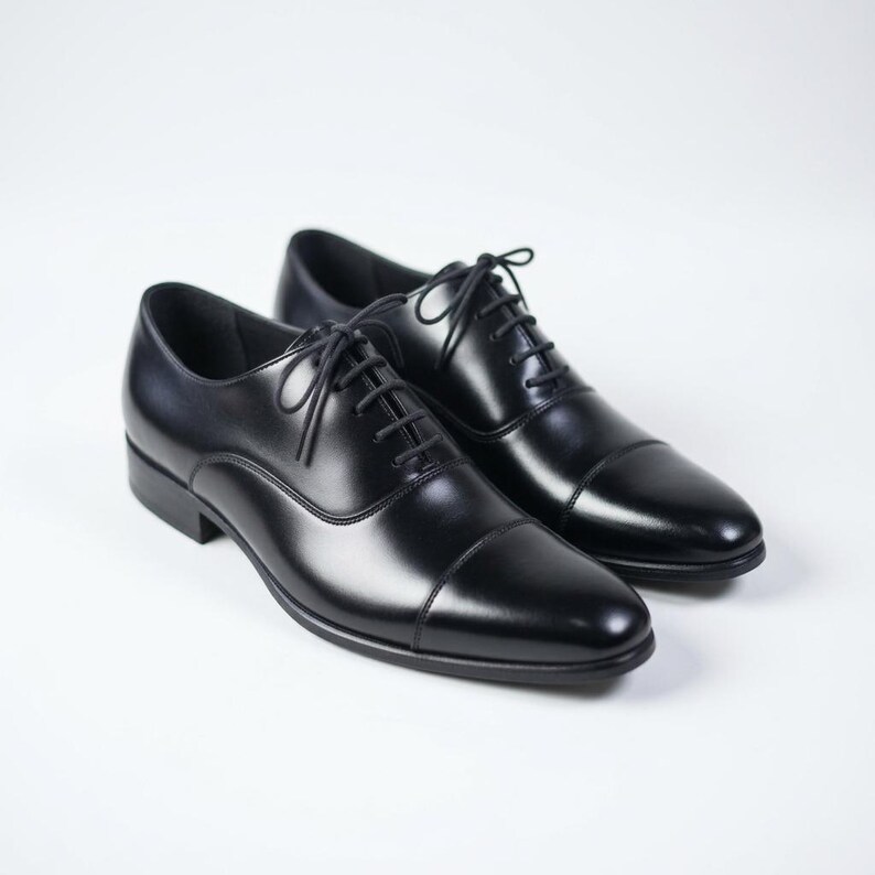 May include: A pair of black leather oxford shoes featuring a cap toe design, a closed lacing system with thin black laces, and a low heel. The shoes have a smooth, polished finish and a rounded toe shape, presenting a clean and structured appearance.
