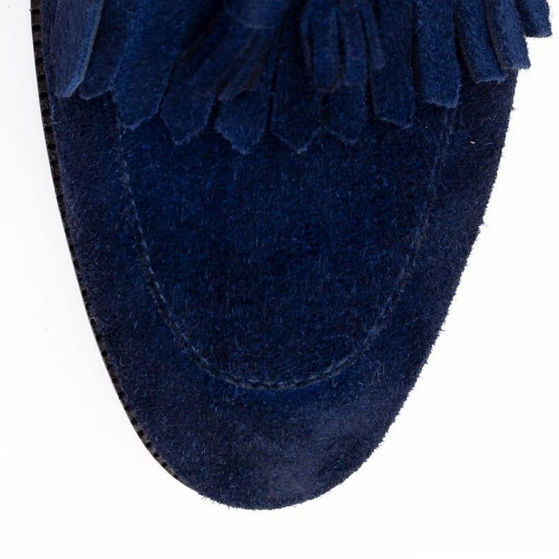 Puede incluir: Primer plano de un mocas&iacute;n de ante azul marino. El zapato presenta una puntera redondeada y un detalle de flecos decorativos en la parte superior. El ante tiene un aspecto suave y texturizado. Las costuras son visibles en los bordes.