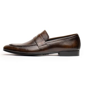 May include: A brown leather loafer shoe with a penny strap. The shoe has a rounded toe and a low heel. The leather has a polished finish and a slight sheen. The shoe is designed for formal or business wear.