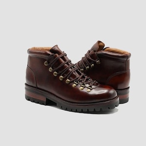 Puede incluir: Un par de botas de senderismo de cuero marrón con cordones marrones oscuros y ojales dorados. Las botas tienen una suela gruesa y acanalada y un cuello acolchado. El cuero tiene un acabado pulido.