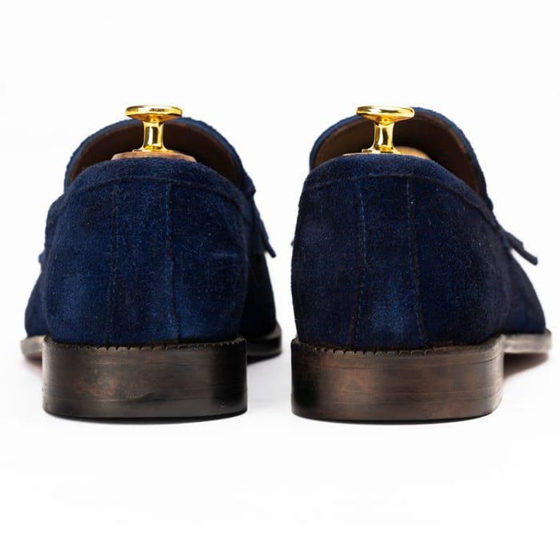 Puede incluir: Un par de mocasines de ante azul marino con suelas marrones oscuras. Los zapatos tienen un dise&ntilde;o cl&aacute;sico, con un detalle de borla y una horma de madera en el interior. El fondo es blanco.