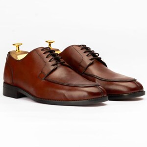 May include: A pair of brown leather dress shoes with black laces. The shoes have a classic design with a rounded toe and a low heel. Shoe trees are inserted in the shoes. The shoes are set against a white background.