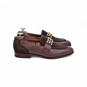 K&ouml;nnte beinhalten: Ein Paar braune Leder-Loafer mit goldfarbenen Schnallen. Die Schuhe haben einen Wildleder-Akzent am Blatt und werden auf h&ouml;lzernen Schuhspannern pr&auml;sentiert. Das Leder hat eine strukturierte Oberfl&auml;che.