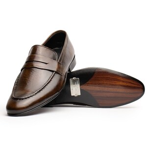 May include: Brown leather penny loafers with a polished finish. The shoes feature a classic design with a strap across the vamp. The sole is a dark brown color with a wood grain pattern. The shoes are displayed on a white background.