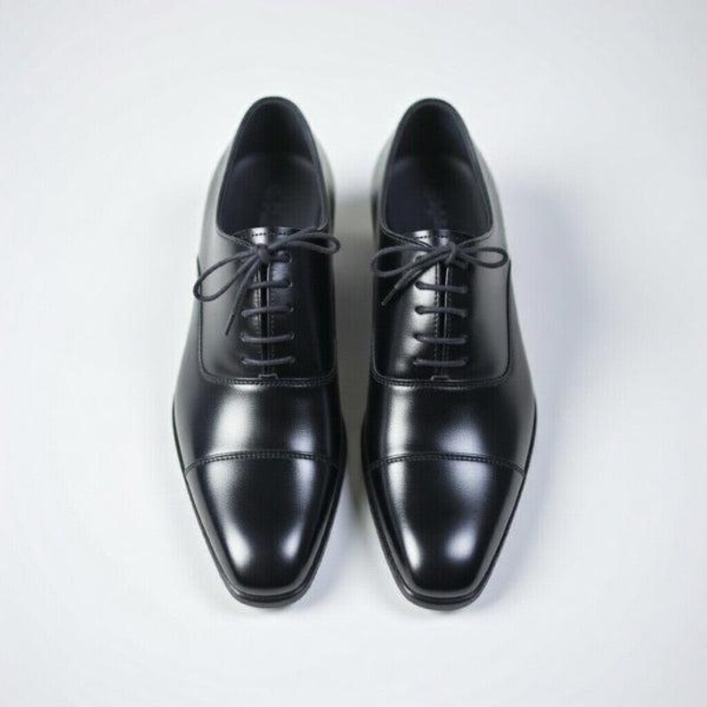 May include: A pair of black leather oxford shoes featuring a classic cap toe design, a smooth polished finish, and thin black laces threaded through five eyelets on each side. The shoes have a structured silhouette with a rounded toe box.