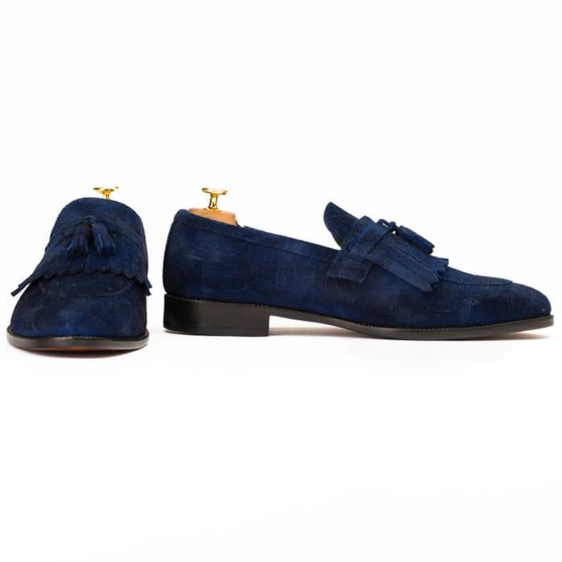 Puede incluir: Un par de mocasines de ante azul marino con detalles de borlas. Estos zapatos sin cordones tienen un dise&ntilde;o cl&aacute;sico con un tac&oacute;n bajo y se muestran sobre un fondo blanco. Los mocasines son una opci&oacute;n de calzado elegante.