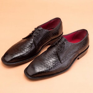 Puede incluir: Un par de zapatos de vestir de cuero negro con un patrón texturizado similar al de un reptil. Los zapatos tienen punta cuadrada, cordones y un forro interior rojo. Los zapatos se exhiben sobre una superficie de color beige claro.