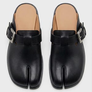 May include: A pair of black leather slip-on shoes featuring a distinct split-toe design. Each shoe includes a wide strap across the top secured with a large, brushed silver-tone metal buckle. The interior footbed is a light tan color.