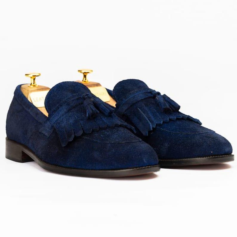 Puede incluir: Un par de mocasines de ante azul marino con detalles de borlas. Los zapatos tienen un dise&ntilde;o cl&aacute;sico con tac&oacute;n bajo y estilo sin cordones. Los mocasines se exhiben sobre hormas de madera sobre un fondo blanco.