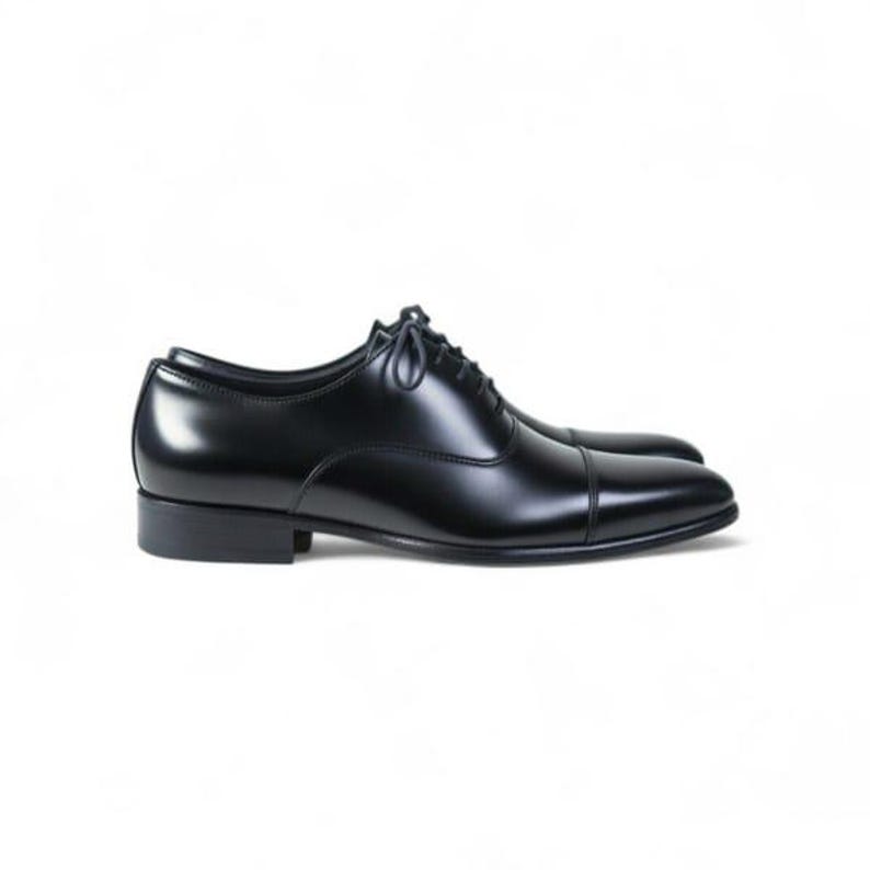 May include: A polished black leather oxford shoe featuring a classic cap toe design, a low stacked heel, and thin black laces. The shoe has a sleek, smooth finish with visible stitching along the toe cap and side panels, set against a plain white background.