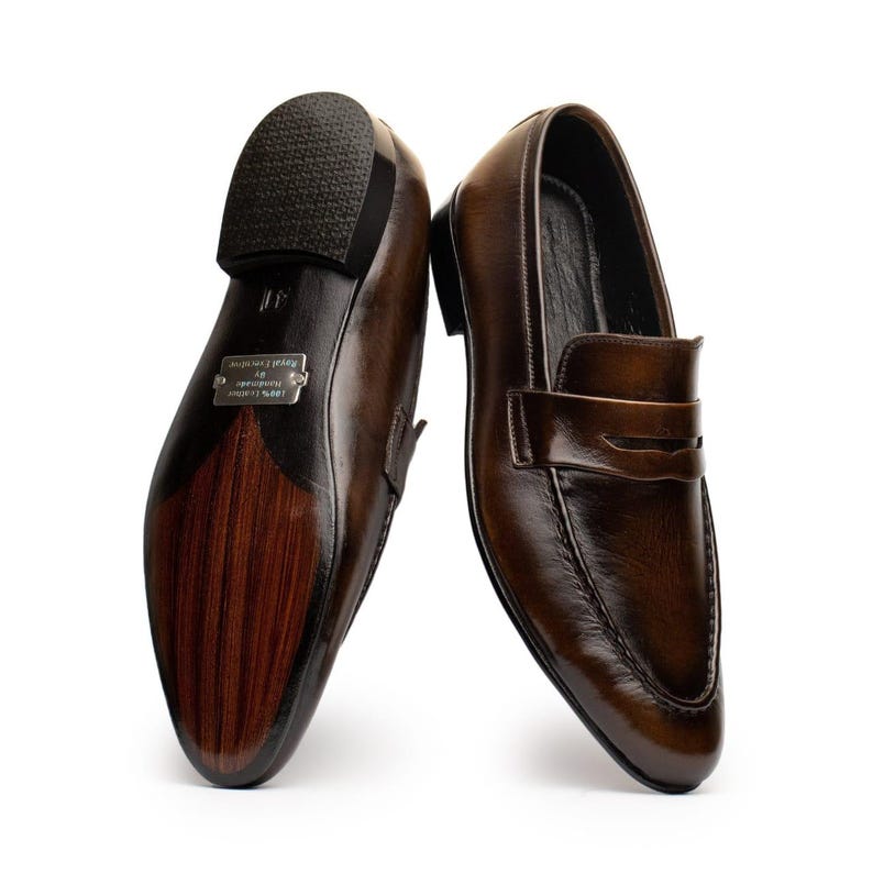 May include: A pair of brown leather loafers with a glossy finish. The shoes have a classic penny loafer design with a rounded toe and a low heel. The soles are dark brown with a textured rubber heel.