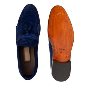 Puede incluir: Un par de mocasines de ante azul marino con detalles de borlas. Un zapato se muestra desde arriba, el otro desde la suela, revelando una suela de madera con la palabra "LEATHER" impresa.