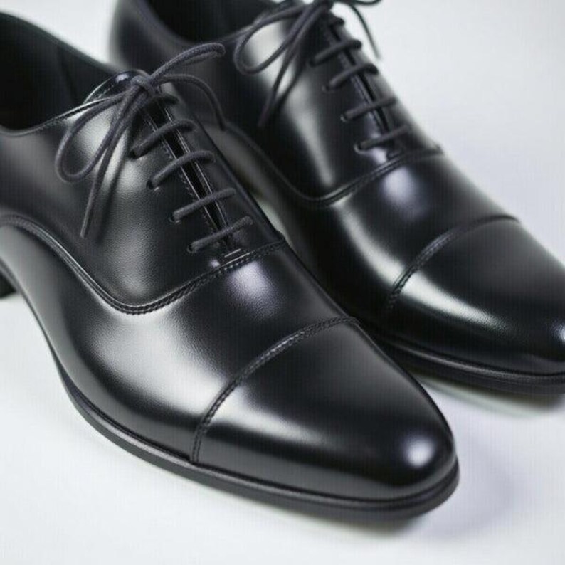 May include: A pair of black leather oxford shoes featuring a smooth, polished finish, a classic cap toe design, and thin black laces threaded through five eyelets on each side. The shoes have a structured silhouette with visible stitching details.