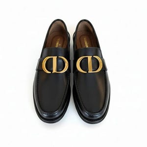 Pode incluir: Um par de mocassins de couro preto com detalhes dourados. Os sapatos apresentam um logotipo 'CD' dourado na tira. Os sapatos são exibidos em um fundo branco.