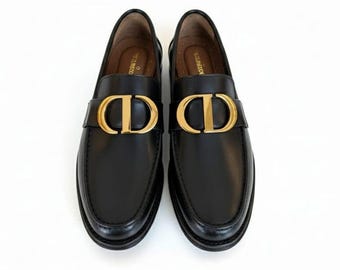 May include: A pair of black leather loafers with gold-colored hardware. The shoes feature a gold-colored 'CD' logo on the strap. The shoes are displayed on a white background.