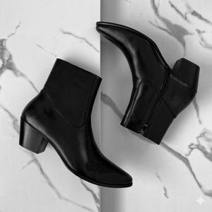 May include: Two black leather ankle boots with pointed toes and block heels. One boot is angled towards the viewer, showcasing the side zipper. The boots are set against a white and gray marble background.