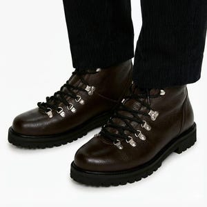 Puede incluir: Botas de senderismo de cuero marrón con cordones negros y ojales metálicos plateados. Las botas tienen una suela negra gruesa y se usan con pantalones de pana negros.