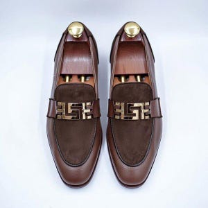 K&ouml;nnte beinhalten: Ein Paar braune Leder-Loafer mit einem dunkelbraunen Wildleder-Vorderblatt und einem auff&auml;lligen goldfarbenen Metallbeschlag auf der Oberseite. Jeder Schuh enth&auml;lt einen Holz-Schuhspanner mit einem goldenen Knopf an der Ferse.