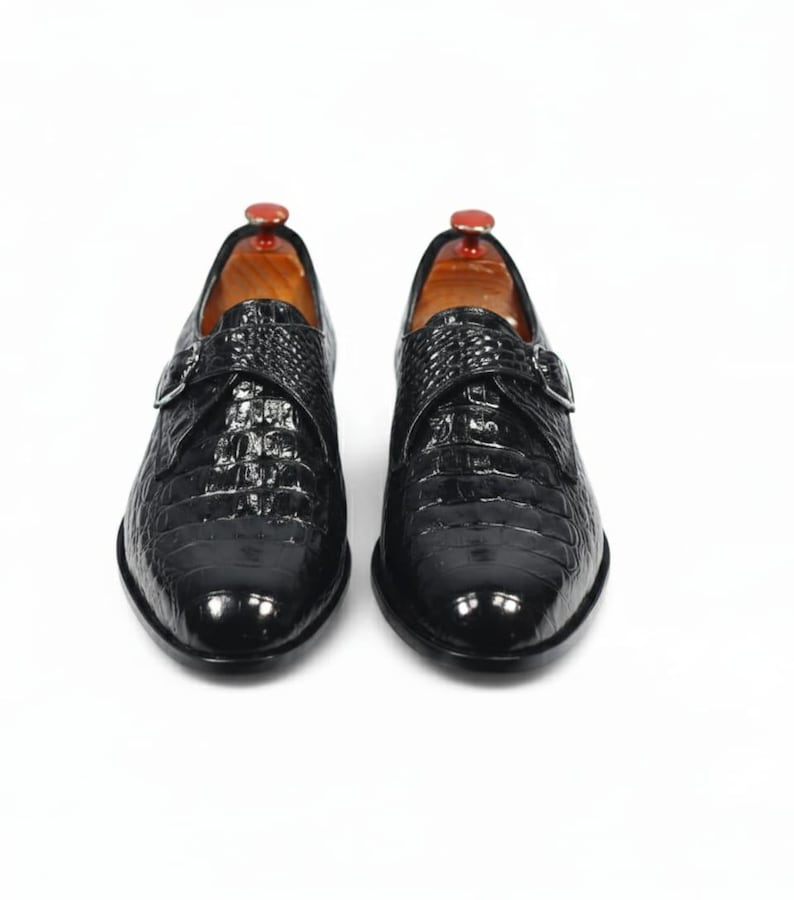 Puede incluir: Un par de zapatos de vestir de cuero negro con textura de piel de cocodrilo. Los zapatos tienen cierre de hebilla y se muestran sobre un fondo blanco. Los zapatos son formales y elegantes.