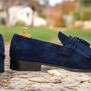 Puede incluir: Un par de mocasines de ante azul marino con detalles de borlas. Los zapatos tienen un tac&oacute;n bajo y se exhiben sobre una superficie de piedra. El fondo est&aacute; borroso, con vegetaci&oacute;n y edificios.