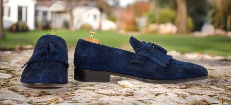 Puede incluir: Un par de mocasines de ante azul marino con detalles de borlas. Los zapatos tienen un tac&oacute;n bajo y se exhiben sobre una superficie de piedra. El fondo est&aacute; borroso, con vegetaci&oacute;n y edificios.